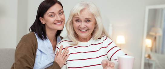 Enthusiastic elder woman and caregiver looking at camera while gathering puzzle